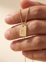 Collar Inicial de Emma Montiel: colgante rectangular con inicial grabada, acabado dorado cálido, sobre cadena tipo box, presentado completo sobre fondo crudo.